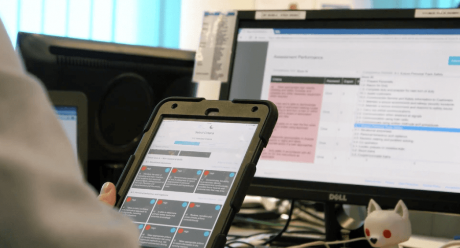 Mobile phone displaying an app, being held by its user as they sit at a desk in front of a PC.