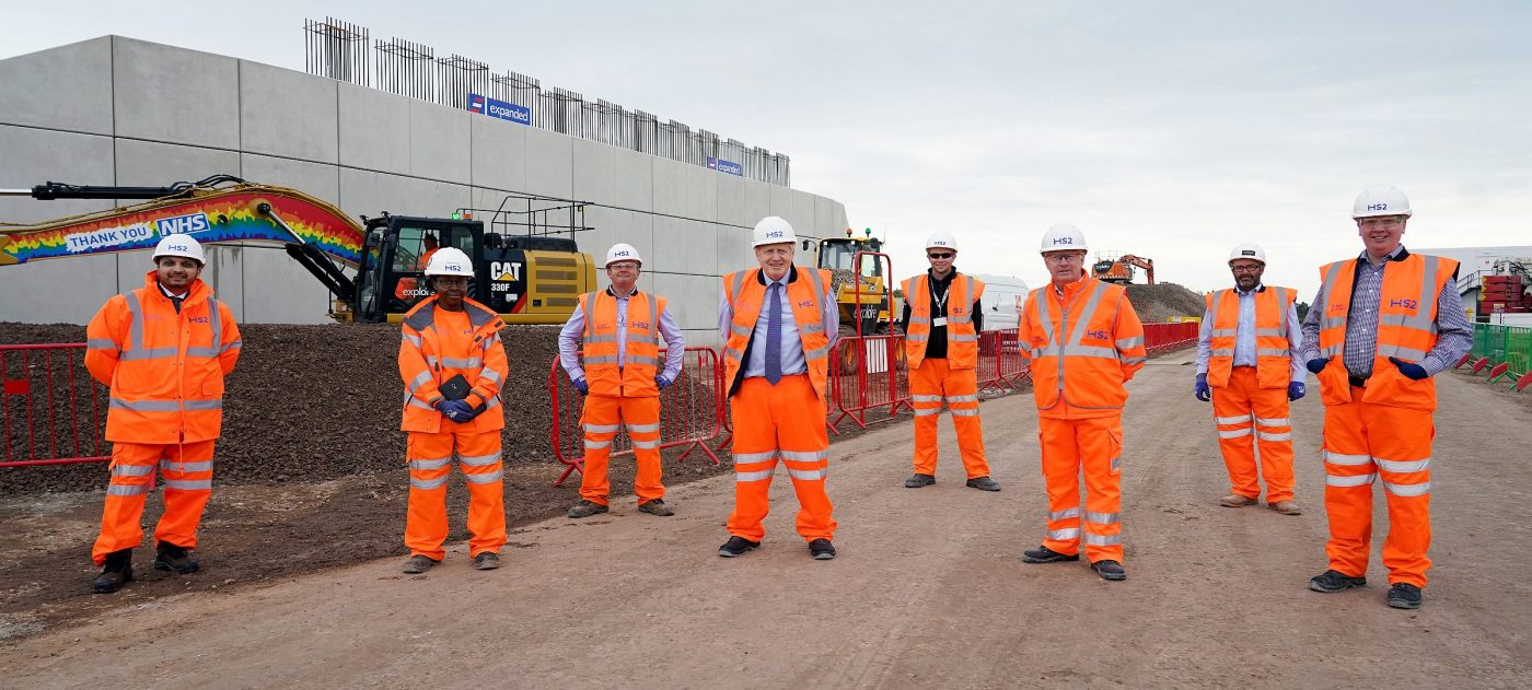 Prime Minister Boris Johnson stands with HS2 workes between two bridge abutments on a construction site.