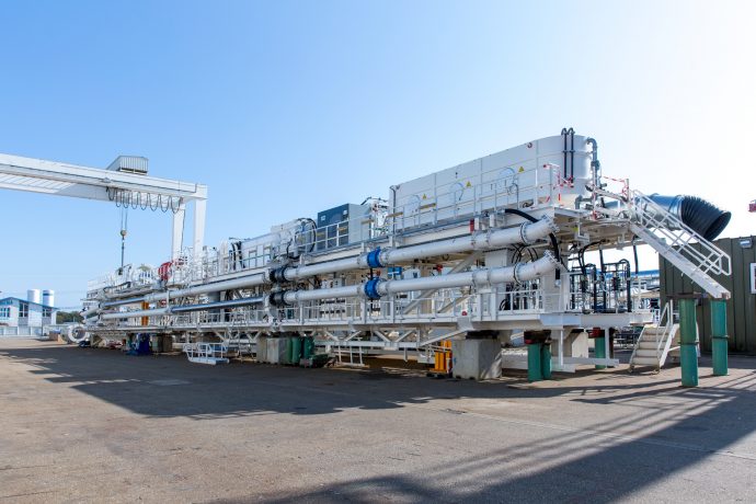 A tunnel boring machine, over 170 metres long sits in a factory yard. Along its side, ducts, pipes and supports are visible.